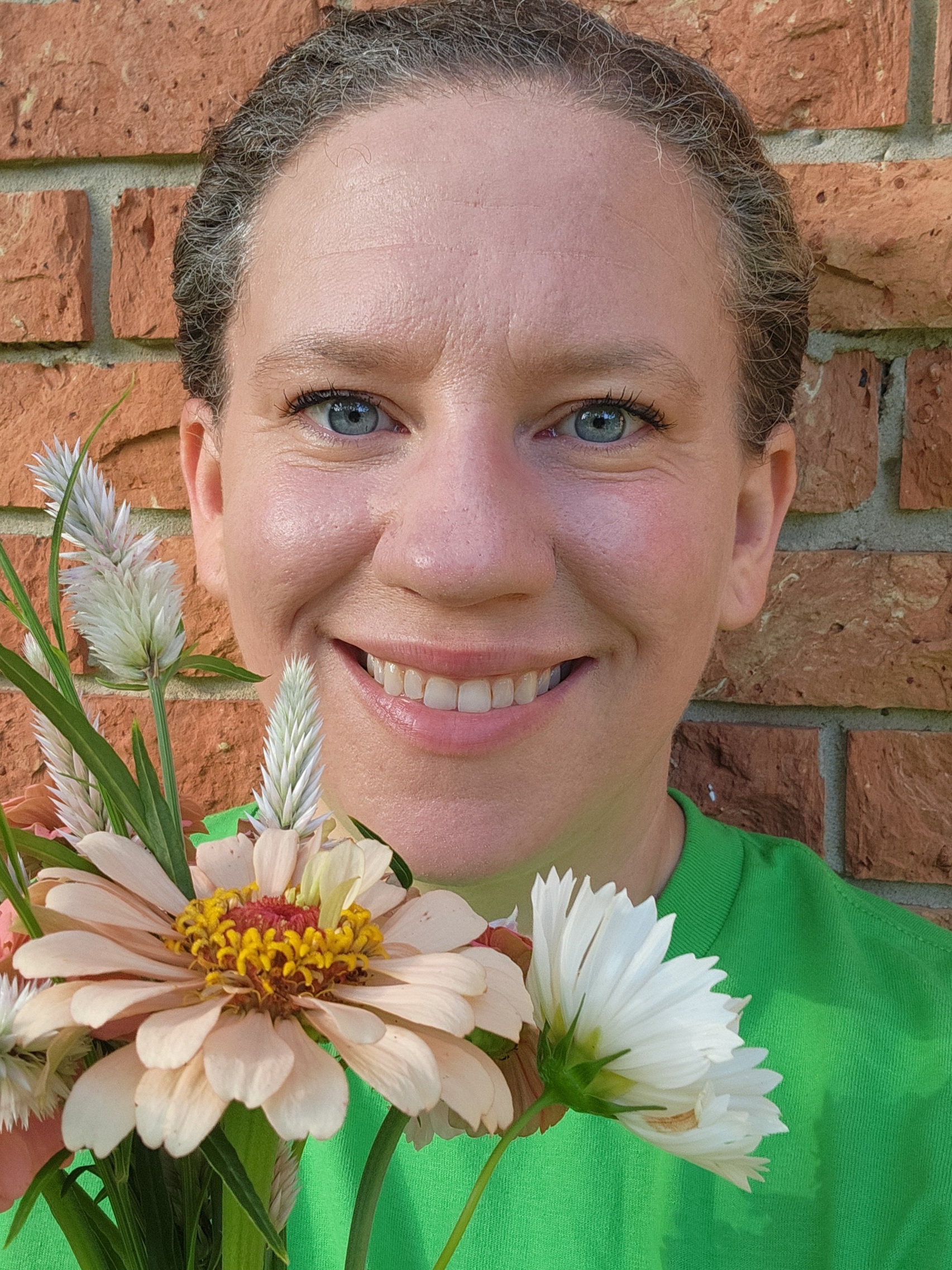 A photo of a dark haired woman holding pink and white flowers with a brick wall in the background.